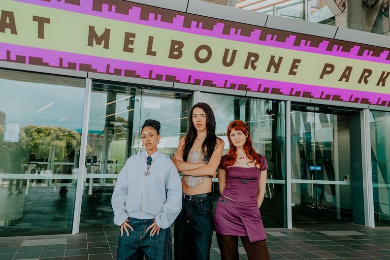Three members of SUPAGLOO pose outside Melbourne Park venue under a bright pink and yellow marquee sign for a promotional photo shoot.